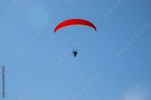 Paramotor (powered paraglider) with red parachute flying in blue sky. Back view with some flare.