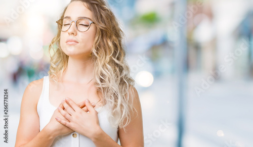 Beautiful young blonde woman wearing glasses over isolated background smiling with hands on chest with closed eyes and grateful gesture on face. Health concept.
