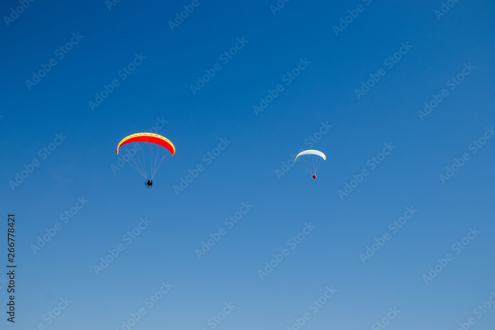 Two paramotors (powered paragliders) with red and yellow parachutes flying in blue sky. Two paramotors (powered paragliders) with red and yellow parachutes flying in blue sky.