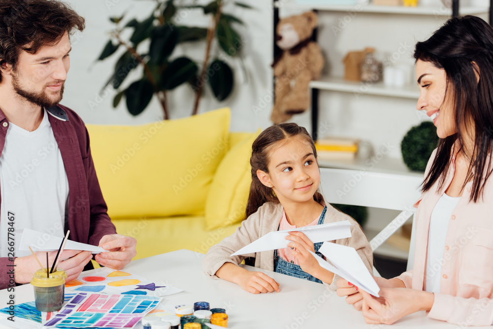 Fototapeta premium happy kid looking at mother holding paper plane while sitting near father