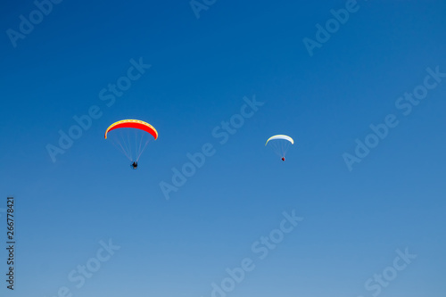 Two paramotors  powered paragliders  with red and yellow parachutes flying in blue sky.