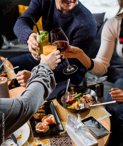 Friends clink glasses at a restaurant during brunch. A man and two women drink wine, fresh orange and green smoothies in a breakfast restaurant. Table with dishes. Close-up. Top view