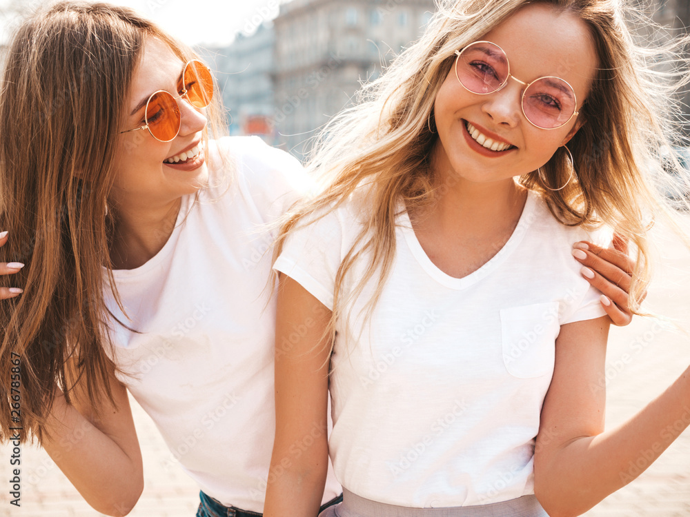 Portrait of two young beautiful blond smiling hipster girls in trendy summer white t-shirt clothes. Sexy carefree women posing on street background. Positive models having fun in sunglasses.Hugging Portrait of two young beautiful blond smiling hipster girls in trendy summer white t-shirt clothes. Sexy carefree women posing on street background. Positive models having fun in sunglasses.Hugging
