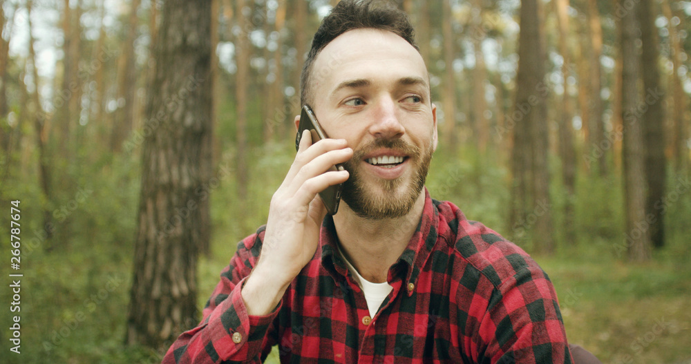 Handsome young bearded man has pleasant talk via smartphone while relaxing in the pine forest Handsome young bearded man has pleasant talk via smartphone while relaxing in the pine forest