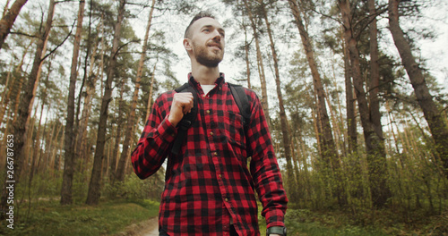 Happy young bearded man dressed in red schecked shirt and backpack walking in the pine forest