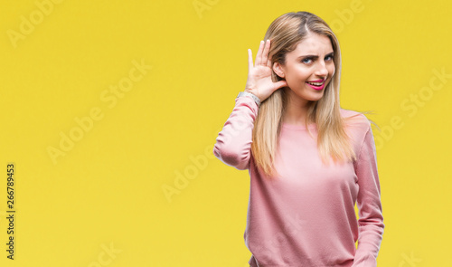 Young beautiful blonde woman wearing pink winter sweater over isolated background smiling with hand over ear listening an hearing to rumor or gossip. Deafness concept.