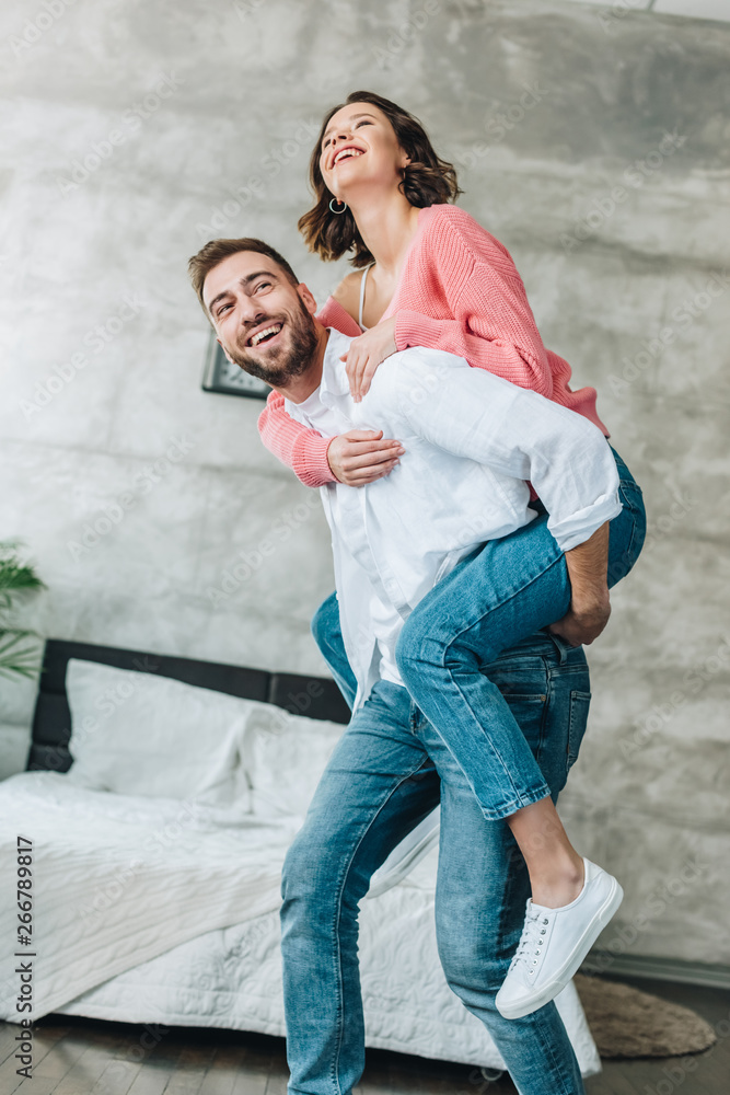 low angle view of happy bearded man piggybacking brunette woman in bedroom