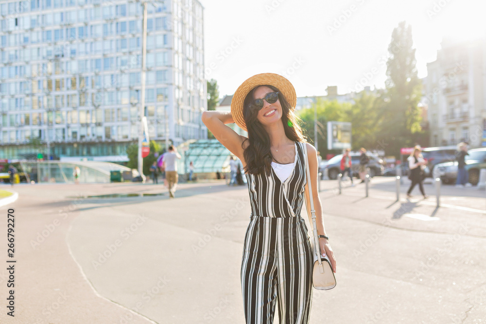 Outside portrait of smiling charming brunette woman wearing stripped suit and straw hat on sunny street in summer weekend