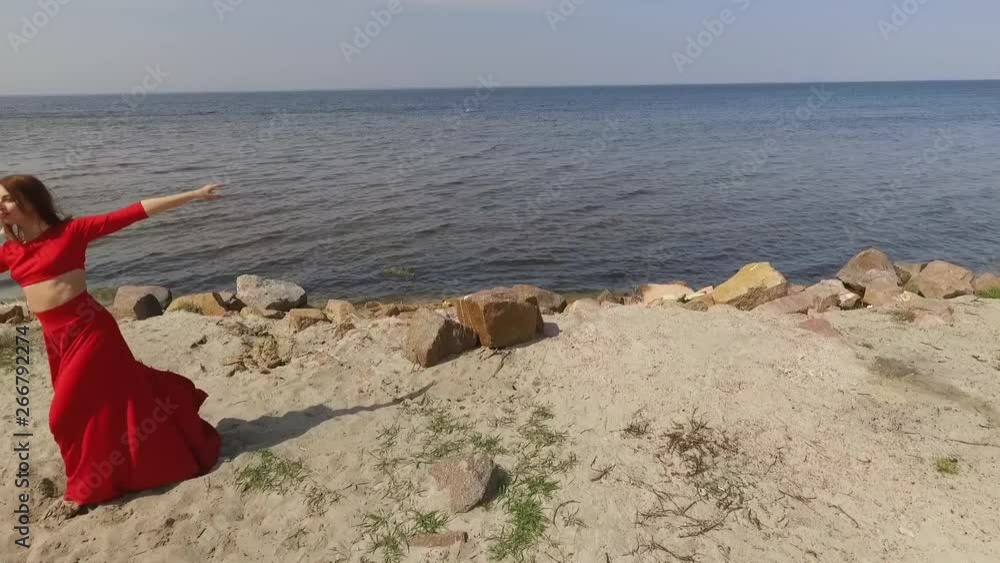 Young graseful woman in red long dress dancing raises hands on the big stone near spectacular view of blue sea water and amazing sky. Contemporary dancer practicing outdoors. Shooting on drone. Slow Young graseful woman in red long dress dancing raises hands on the big stone near spectacular view of blue sea water and amazing sky. Contemporary dancer practicing outdoors. Shooting on drone. Slow