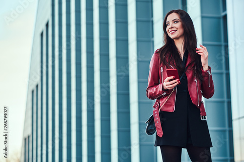 Beautiful smiling woman in red jacket is standing next to glass building with smartphone in her hands.