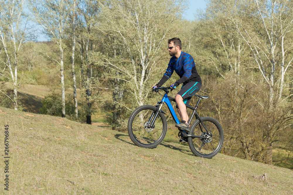 Cyclist in shorts and jersey on a modern carbon hardtail bike with an air suspension fork rides off-road on green hills near the forest	
