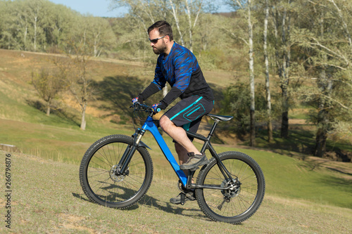 Cyclist in shorts and jersey on a modern carbon hardtail bike with an air suspension fork rides off-road on green hills near the forest 