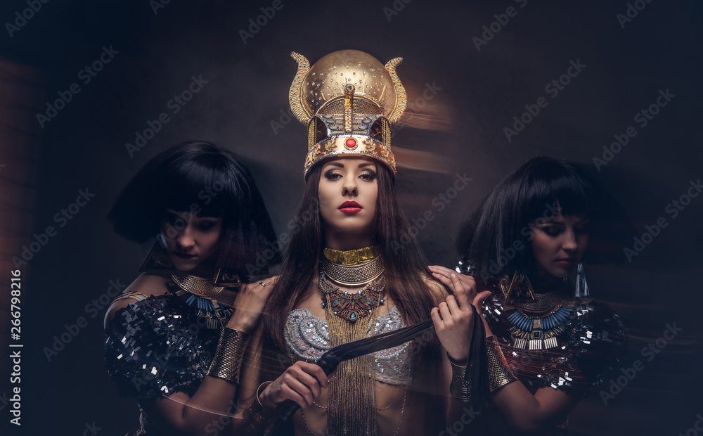 Fototapeta premium Portrait of haughty Egyptian queen in an ancient pharaoh costume with two concubines. Isolated on a dark background.
