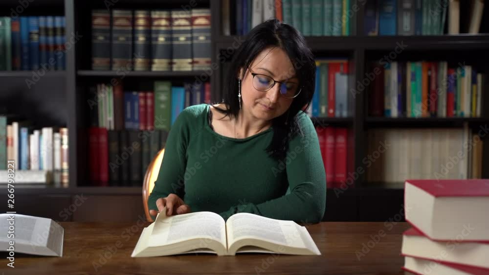 Beautiful young librarian woman sitting at wooden table and reading book in local library