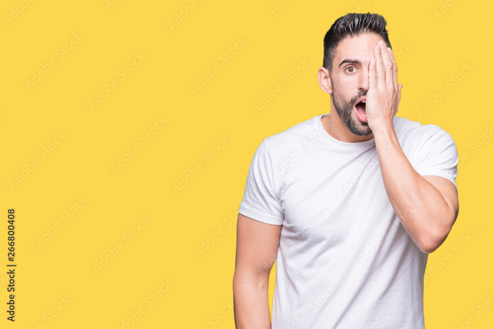Young man wearing casual white t-shirt over isolated background covering one eye with hand with confident smile on face and surprise emotion. Young man wearing casual white t-shirt over isolated background covering one eye with hand with confident smile on face and surprise emotion.