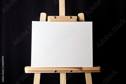 White blank cotton canvas stands on a wooden artistic easel on black curtain background. Horizontal rectangular mockup canvas wrapped on stretcher bar