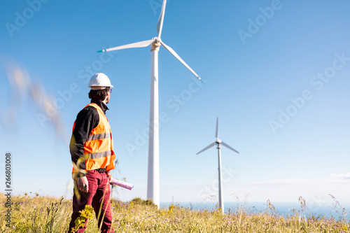 Giovane ingegnere con gilet ad alta visibilità, casco bianco e progetto in mano, sta controllando l'impianto eolico formato da turbine a vento in montagna. Concetto di manutenzione.