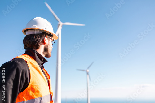 Giovane ingegnere con gilet ad alta visibilità, casco bianco e progetto in mano, sta controllando l'impianto eolico formato da turbine a vento in montagna. Concetto di manutenzione.