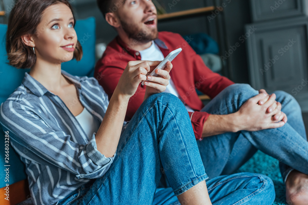 selective focus of attractive woman holding smartphone near surprised man selective focus of attractive woman holding smartphone near surprised man