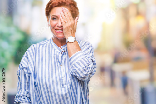 Atrractive senior caucasian redhead woman over isolated background covering one eye with hand with confident smile on face and surprise emotion.