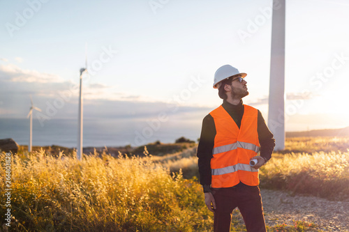 Giovane Ingegnere Aerospaziale con caschetto bianco e gilet arancione, sta controllando il progetto sul foglio di una turbina di un impianto eolico in montagna. Concetto ingegneria ed energia pulita © Polonio Video