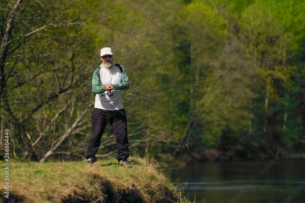 Fisherman throwing the bait into river standing on river bank in sunny day with green forest in background Fisherman throwing the bait into river standing on river bank in sunny day with green forest in background