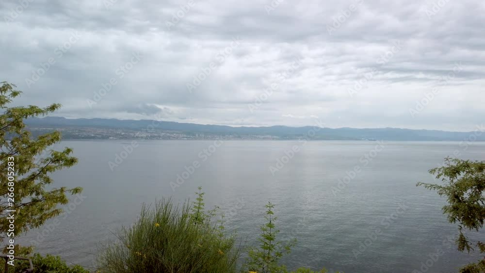 Panning view across calm, peaceful Lovran, Croatia coastline waterfront. Still ocean seascape & coastal tree foliage in foreground. Panning view across calm, peaceful Lovran, Croatia coastline waterfront. Still ocean seascape & coastal tree foliage in foreground.