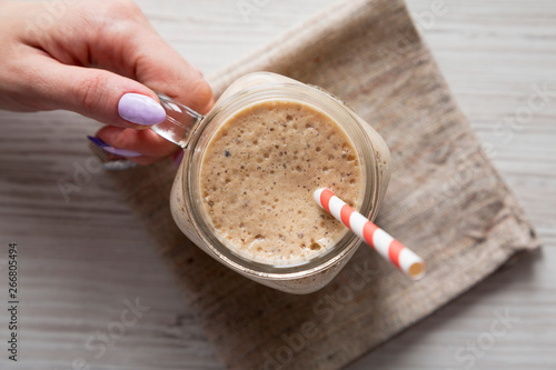 Woman's hand holding a glass jar filled with banana, kiwi, apple smoothie over white wooden surface, top view. Flat lay, from above, overhead.