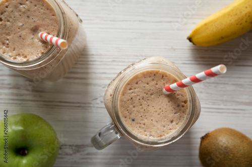 Glass jars filled with banana, kiwi, apple smoothie on a white wooden background. From above, overhead, top view.