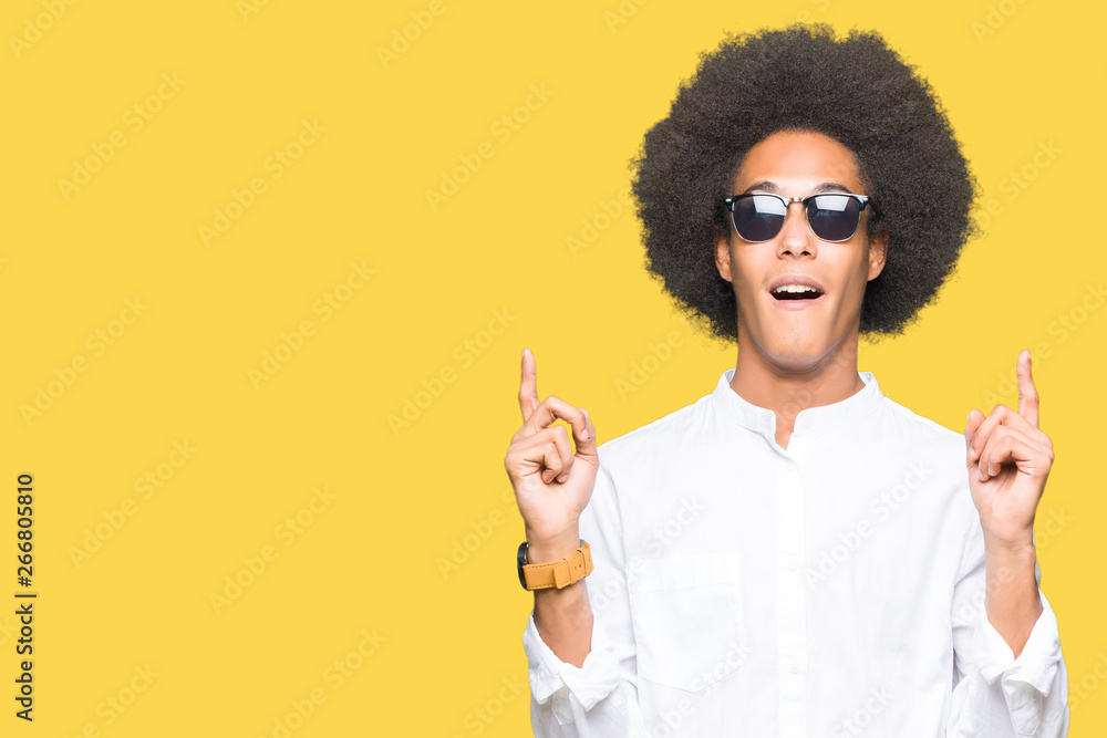 Young african american man with afro hair wearing sunglasses amazed and surprised looking up and pointing with fingers and raised arms. Young african american man with afro hair wearing sunglasses amazed and surprised looking up and pointing with fingers and raised arms.