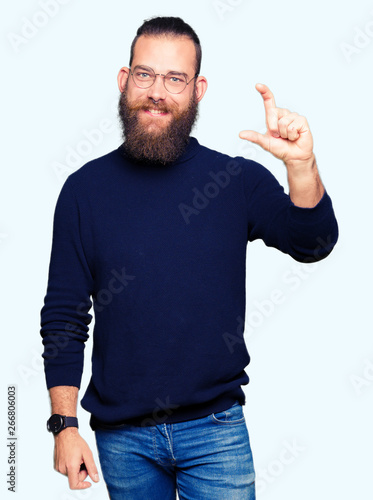 Young blond man wearing glasses and turtleneck sweater smiling and confident gesturing with hand doing size sign with fingers while looking and the camera. Measure concept.