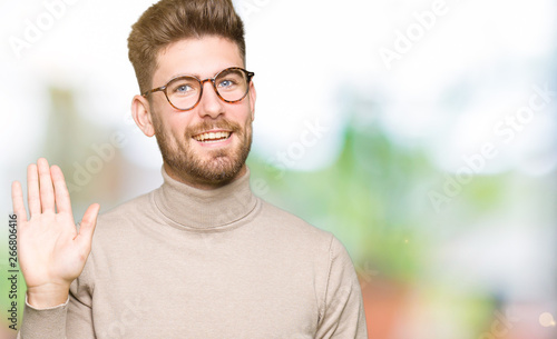 Young handsome business man wearing glasses Waiving saying hello happy and smiling, friendly welcome gesture