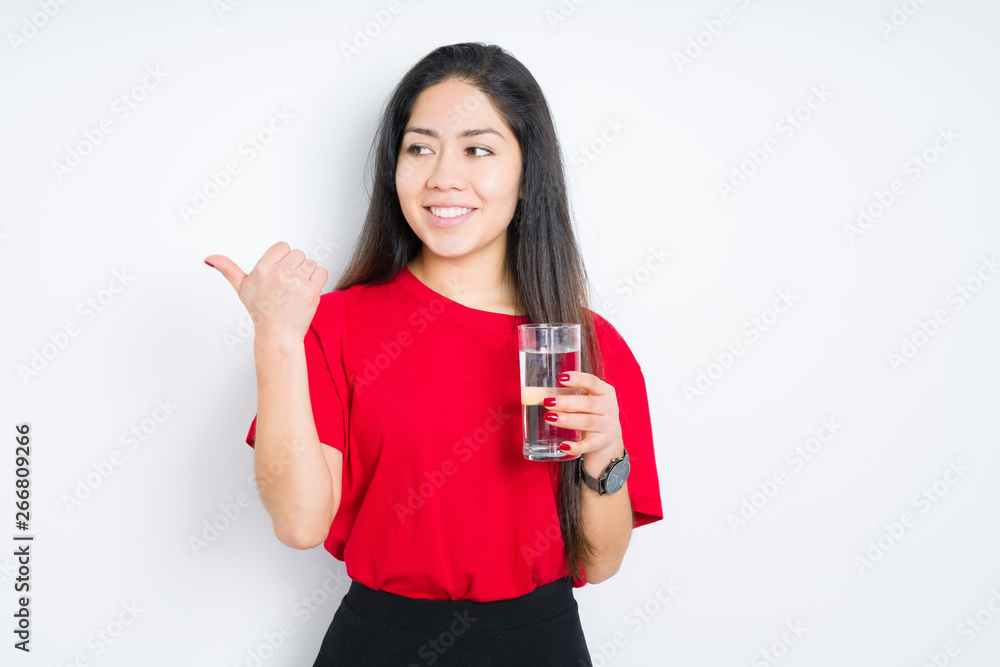 Young brunette woman drinking a glass of water over isolated background pointing and showing with thumb up to the side with happy face smiling