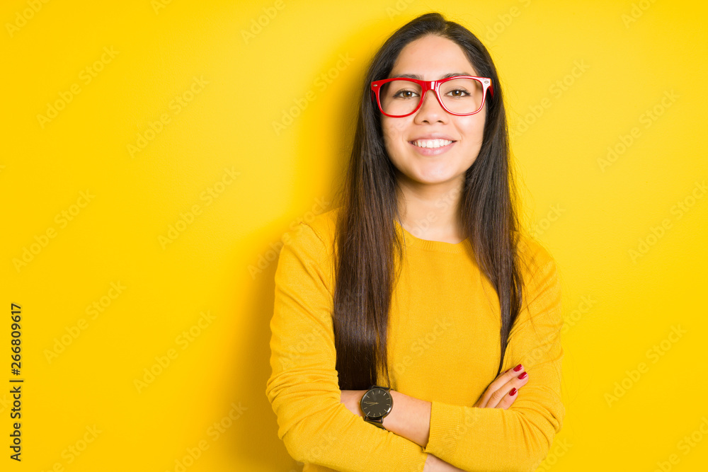 Beautiful brunette woman wearing red glasses over yellow isolated background happy face smiling with crossed arms looking at the camera. Positive person.