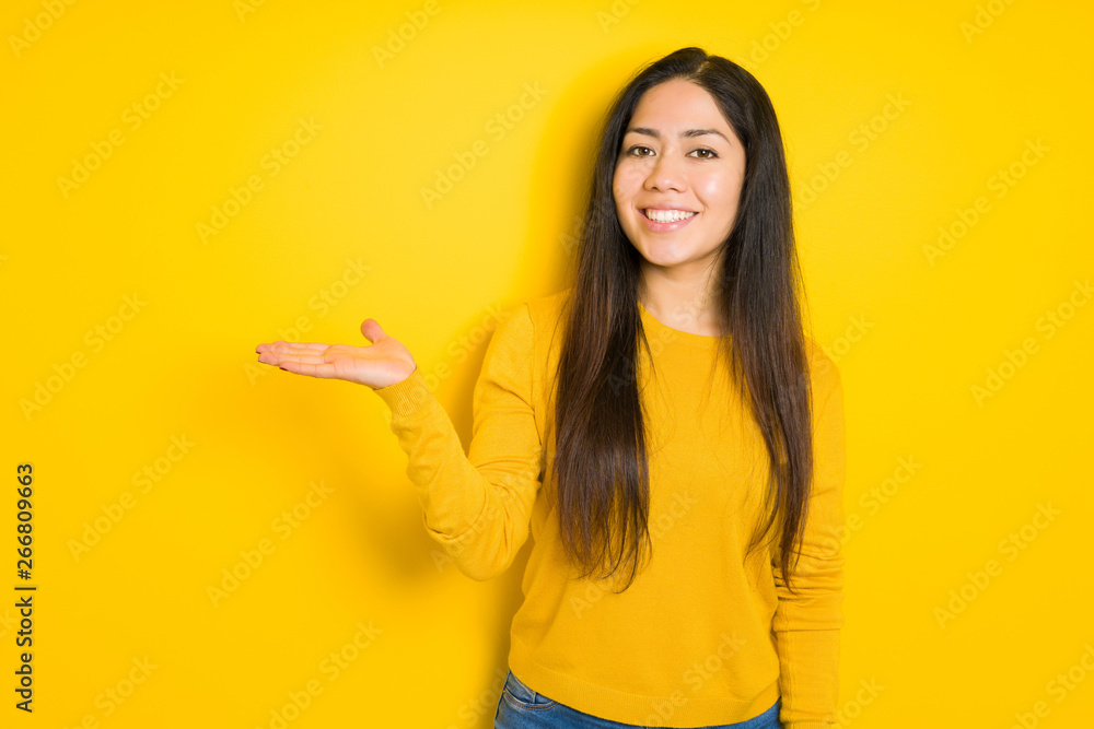 Beautiful brunette woman over yellow isolated background smiling cheerful presenting and pointing with palm of hand looking at the camera. Beautiful brunette woman over yellow isolated background smiling cheerful presenting and pointing with palm of hand looking at the camera.