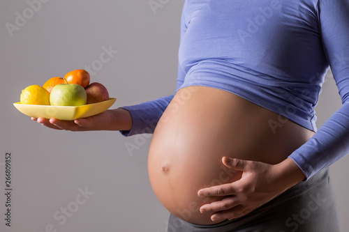 Image of close up stomach of pregnant woman holding fruit on gray background.