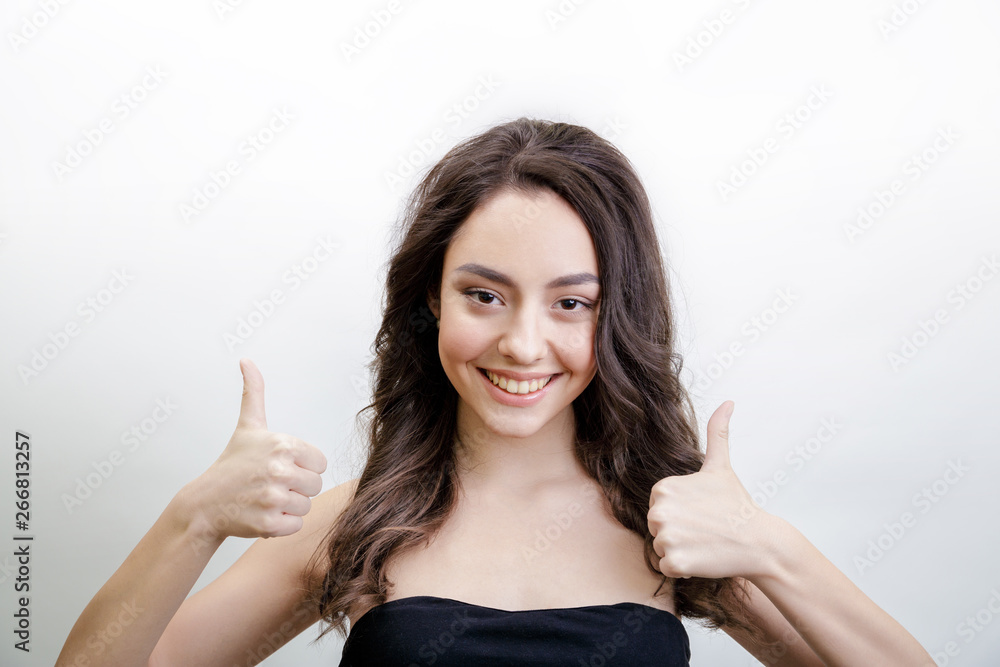 Happy cheerful young woman wearing black strapless dress. Girl has long brown hair with curls. She shows a gesture thumbs up. Woman rejoices positive news or received the necessary credits. Studio. Happy cheerful young woman wearing black strapless dress. Girl has long brown hair with curls. She shows a gesture thumbs up. Woman rejoices positive news or received the necessary credits. Studio.