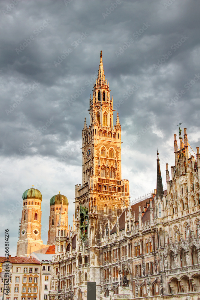 Tall spire of neo-Gothic town hall building Neues Rathaus and towers of Frauenkirche cathedral in historic main square Marienplatz in morning orange sunlight, Altstadt Munchen Bayern Germany Europe