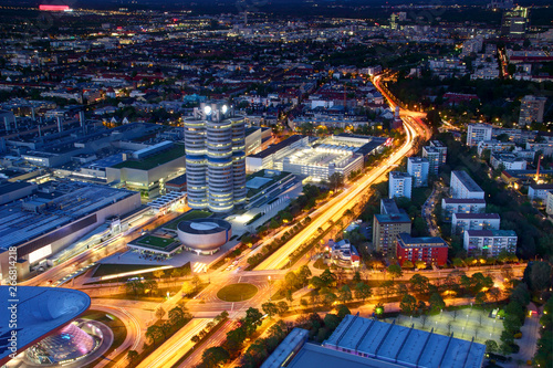 Modern European aerial cityscape in blue hour with broad circle road intersection, commercial, office and industrial buildings in outskirts lit by street and car lights, Munchen Bayern Germany Europe