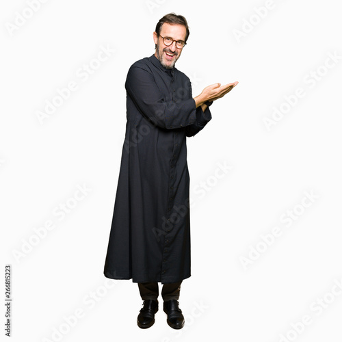 Middle age priest man wearing catholic robe Pointing to the side with hand and open palm, presenting ad smiling happy and confident