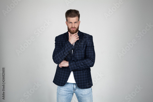 A beautiful sexy bearded man in a jacket holds his hand at the chin and looks thoughtfully at the camera. he stands in front of the white background © Roman