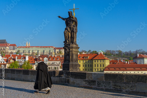 Photography Bishop or priest walk near statue of Saint John the Baptist on pedestal and balustrades of Charles Bridge over Vltava river, and background of Praha Castle in Prague, Czech Republic in sunny day