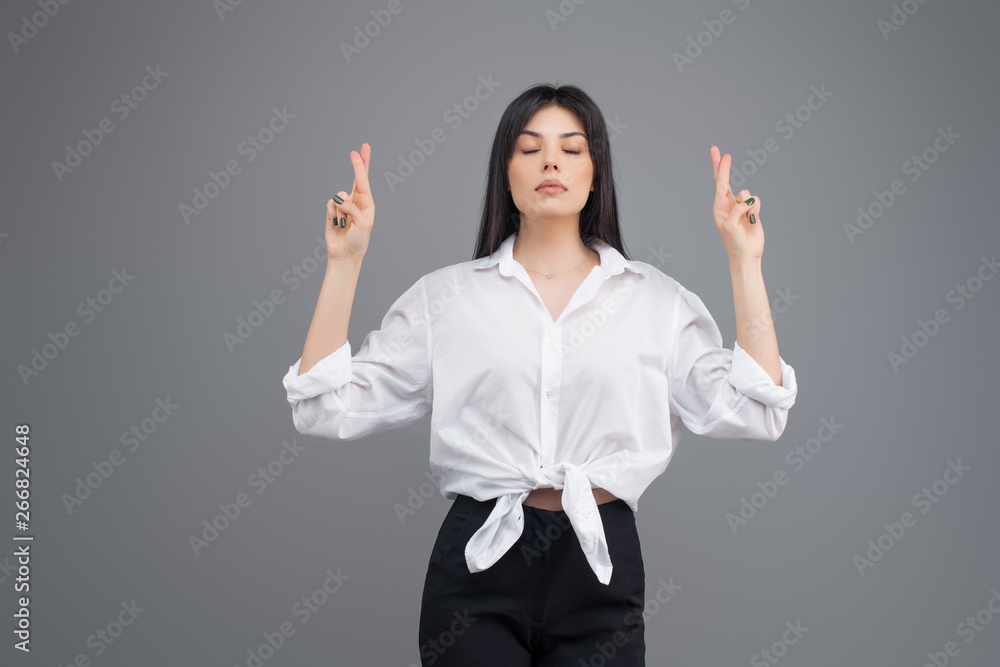 Young business woman praying and hoping for good luck with fingers crossed isolated over grey background