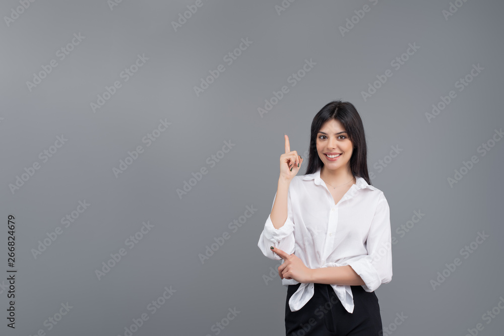Smiling young business woman in white shirt pointing at copy space with her finger isolated over grey background