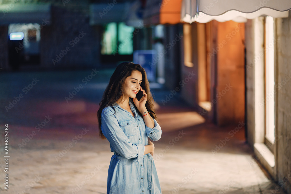 Fototapeta premium A beautiful young girl in a blue dress standing in a night city with phone