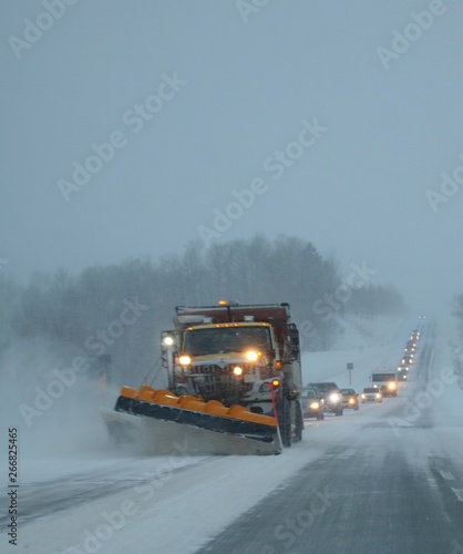 Snowplow clearing away drifting snow on Ontario highway in January 2019 at dusk with long line of vehicles following behind