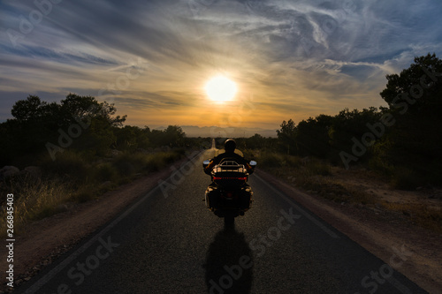 A heavy motorcycle with red lights driving a narrow and completely straight road into the sunset on the horizon with mountains.