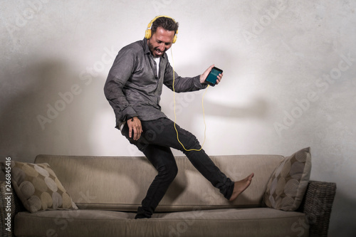 attractive man listening online music with headphones and mobile phone playing air guitar singing song and dancing carefree jumping at living room sofa couch