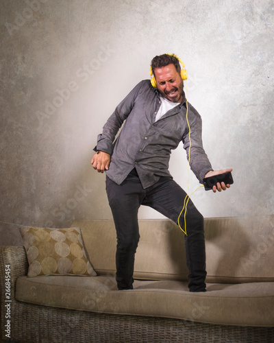 attractive man listening online music with headphones and mobile phone playing air guitar singing song and dancing carefree jumping at living room sofa couch