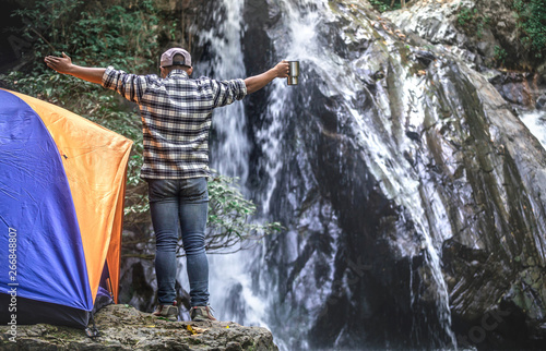 Happy traveler standing with holding coffee cup on rock in front at waterfall, camping concept.